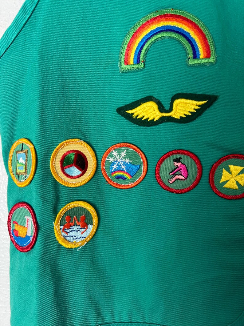 May include: Green fabric with various colorful embroidered patches. Patches include a rainbow, wings, and circular designs with images of a snowflake, a person, and other symbols. The fabric has a pocket.