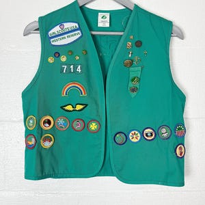 May include: A green Girl Scouts USA vest with various patches and pins. The vest has the text "GIRL SCOUTS USA WESTERN RESERVE" and the number "714". Patches include a rainbow, wings, and various circular designs.
