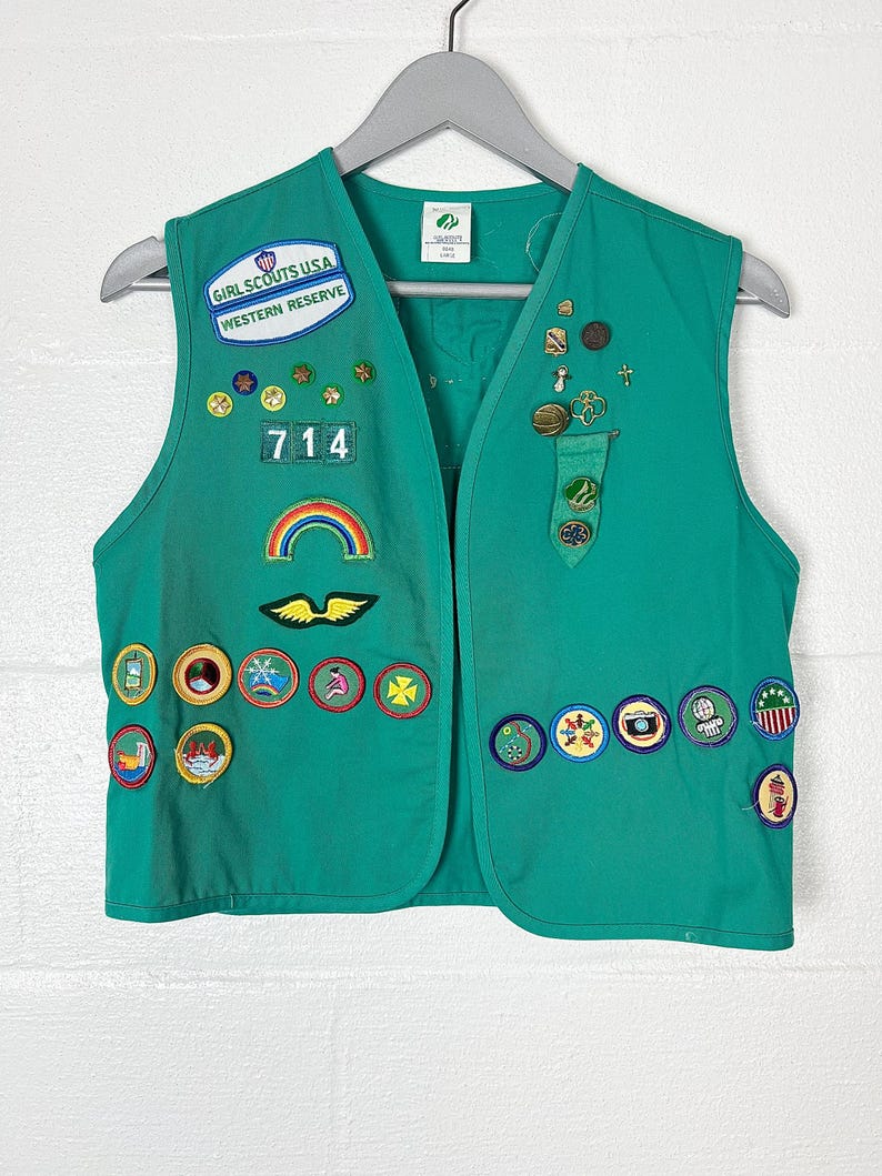May include: A green Girl Scouts USA vest with various patches and pins. The vest has the text "GIRL SCOUTS USA WESTERN RESERVE" and the number "714". Patches include a rainbow, wings, and various circular designs.