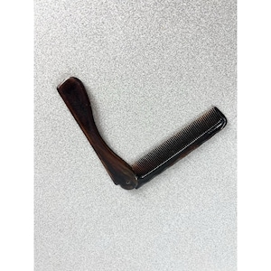 May include: A folding comb with a dark brown, marbled, horn-like finish. The object features a long, curved handle attached to a rectangular section with fine, closely spaced teeth. The two parts are connected by a small metal pivot pin.