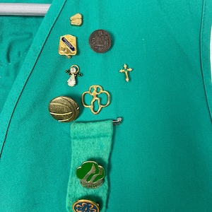 May include: A green vest adorned with various pins, including a gold cross, a Girl Scout emblem, and a pin with the text "Family of God." The vest also features a felt tie with Girl Scout pins.