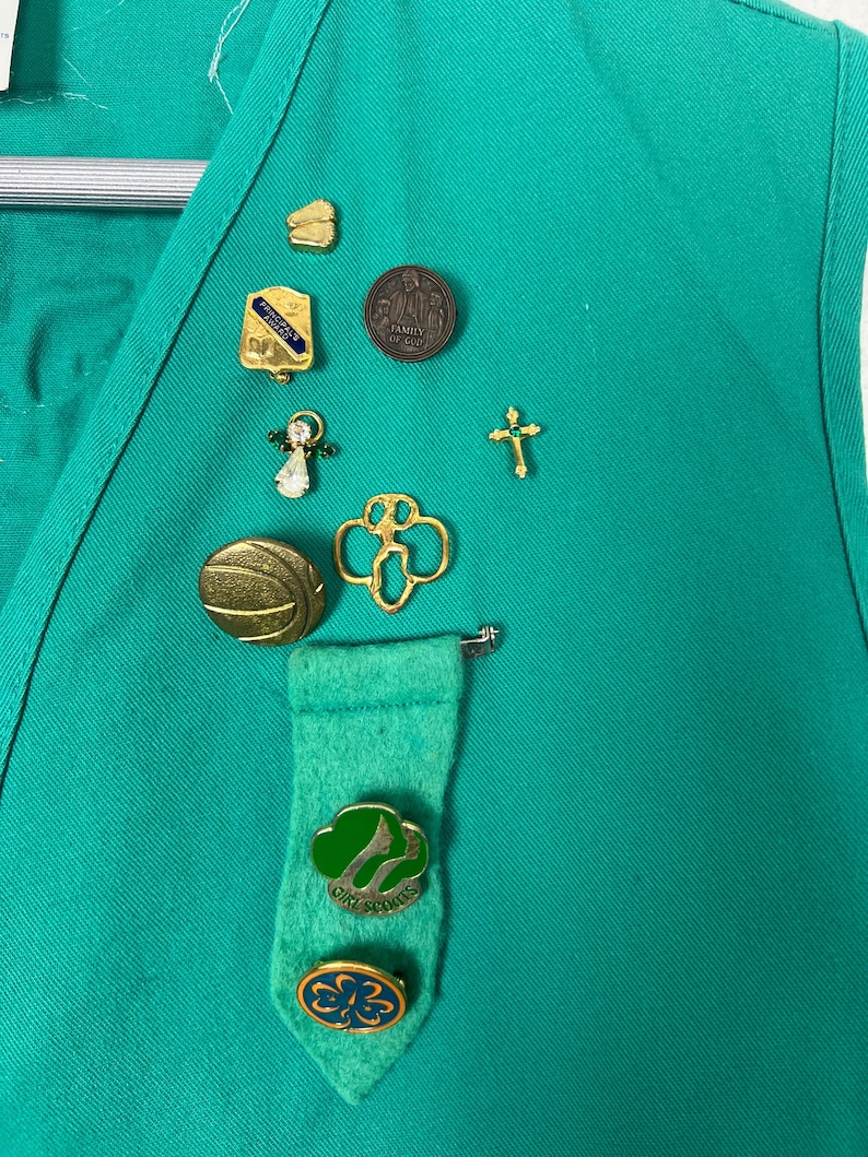 May include: A green vest adorned with various pins, including a gold cross, a Girl Scout emblem, and a pin with the text "Family of God." The vest also features a felt tie with Girl Scout pins.