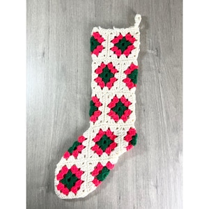 May include: A Christmas stocking made of crocheted granny squares in a repeating pattern of cream, dark green, and bright pink. The stocking features a classic boot shape with a hanging loop at the top corner, constructed from joined squares.