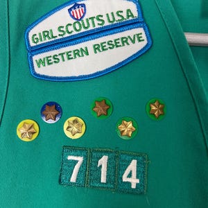 May include: Close-up of a green Girl Scout uniform with patches and pins. Patches include "Girl Scouts USA Western Reserve" and a number "714". Gold star pins and a rainbow patch are also visible.