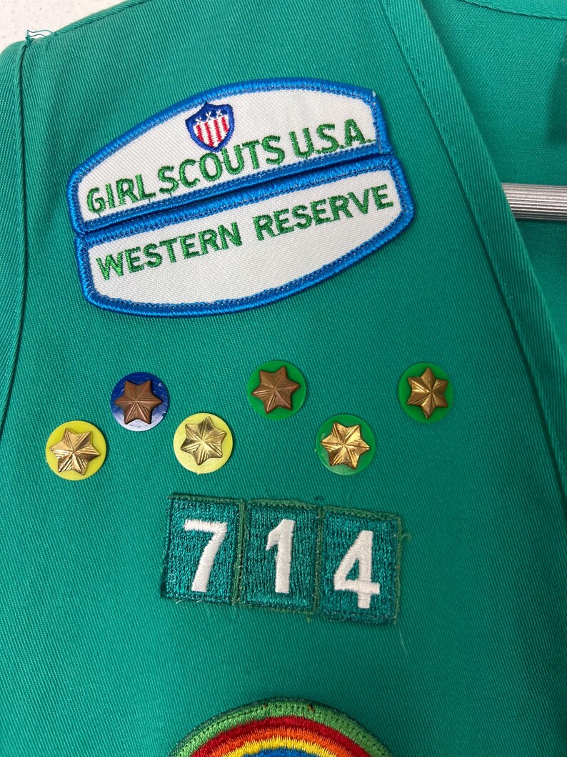 May include: Close-up of a green Girl Scout uniform with patches and pins. Patches include "Girl Scouts USA Western Reserve" and a number "714". Gold star pins and a rainbow patch are also visible.