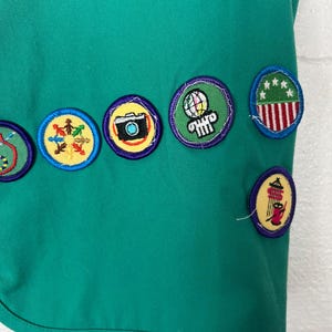 May include: Close-up of a teal green fabric with several embroidered patches. The patches are round with various designs, including a camera, a globe, and a design with red and white stripes. The patches are outlined in yellow and purple.