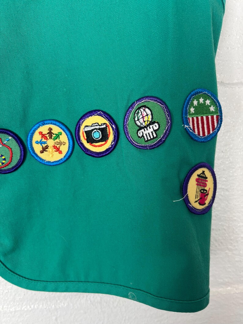 May include: Close-up of a teal green fabric with several embroidered patches. The patches are round with various designs, including a camera, a globe, and a design with red and white stripes. The patches are outlined in yellow and purple.