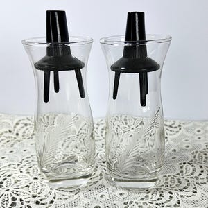 May include: Two clear glass vases with black stoppers. The vases have a flared shape and a decorative etched leaf design. The stoppers have a cylindrical top and a base with three legs. The vases are on a white lace surface.