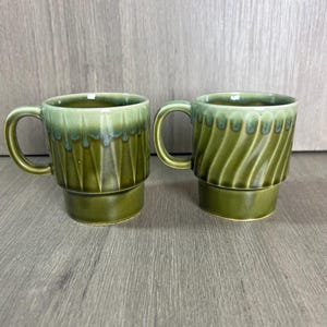 May include: Two ceramic mugs with a glossy olive green glaze and a lighter green drip effect at the rim. The left mug features a vertical geometric pattern, while the right mug has a diagonal ribbed texture. Both have a smooth, solid base.