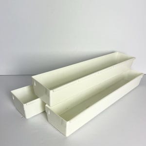 May include: Three off-white rectangular storage bins of varying sizes are stacked. The bins are empty and have a simple, clean design. The bins are made of a smooth, matte material and are arranged against a white background.