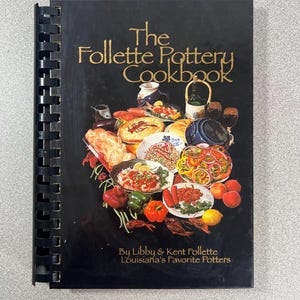 May include: A black spiral-bound cookbook titled "The Follette Pottery Cookbook" with gold lettering. The cover features a colorful collage of food dishes. The text "By Libby & Kent Follette Louisiana's Favorite Potters" is at the bottom.