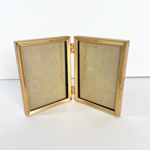 May include: A hinged, gold-colored metal picture frame, open to display two photo spaces. The frame has a textured border and a simple, elegant design. The interior backing is a light brown color.