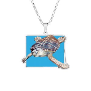 Op de afbeelding: Een zilverkleurige metalen ketting met een rechthoekige hanger met afgeronde hoeken. De hanger toont een gedetailleerde illustratie van een zeeschildpad zwemmend tegen een felblauwe achtergrond, hangend aan een zilveren boxketting.