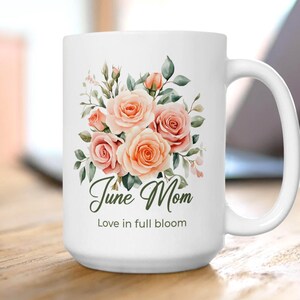 May include: White ceramic mug featuring a floral design with peach and pink roses, green leaves, and the text "June Mom" and "Love in full bloom". The mug has a large handle and is likely used for hot beverages.