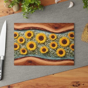 Peut inclure: Une planche &agrave; d&eacute;couper rectangulaire pr&eacute;sentant une bande centrale de tournesols jaunes et de petites fleurs blanches incrust&eacute;es dans une r&eacute;sine bleu clair, bord&eacute;e de deux sections en bois naturel avec des veines et des bords sombres.