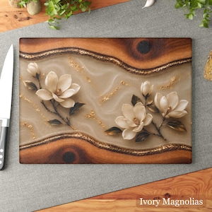 K&ouml;nnte beinhalten: Rechteckiges Holzschneidebrett mit hellbeiger Mitte und dekorativem Rand. Die Mitte zeigt elfenbeinfarbene Magnolienbl&uuml;ten und goldene Akzente. Der Text "Ivory Magnolias" befindet sich unten.