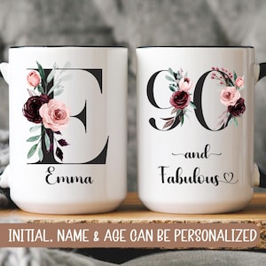 May include: Two white ceramic mugs with black handles. The mug on the left has a black letter "E" with floral designs and the name "Emma" printed below. The mug on the right has the number "90" with floral designs and the text "and Fabulous" printed below.