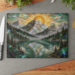 Könnte beinhalten: Ein rechteckiges Glasschneidebrett mit einer Landschaftsillustration im Buntglasstil, die ein Gebirge, eine Sonne, einen Kiefernwald und einen See mit Spiegelung in schillernden Grün-, Blau- und Goldtönen darstellt.