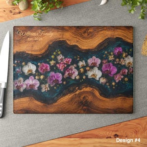 May include: A rectangular wooden cutting board featuring a central river-like inlay of dark blue resin embedded with various white, pink, and purple orchid flowers. The text William Family Est. 2026 is engraved in the upper left corner of the wood.