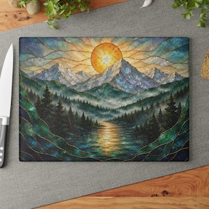 Könnte beinhalten: Ein rechteckiges Glasschneidebrett mit einer Illustration im Buntglasstil, die ein Gebirge, einen Kiefernwald und einen Fluss zeigt, in dem sich eine goldene Sonne spiegelt. Das Design verwendet dunkelblaue, grüne und gelbe Farbtöne mit Goldlinien.