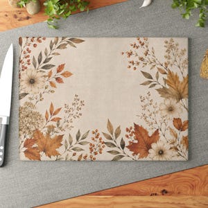 May include: A rectangular glass cutting board featuring a beige background decorated with a botanical print of autumn leaves, berries, and flowers in shades of brown, orange, and cream. A silver kitchen knife is positioned to the left side.