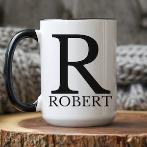 May include: White ceramic mug with a black rim and handle. The mug has a large black letter "R" and the name "ROBERT" printed on the front.