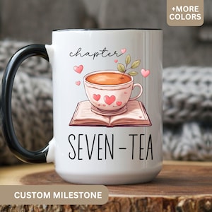 May include: White ceramic mug with a black handle and rim. The mug features a whimsical illustration of a teacup on a book, with the words "chapter SEVEN-TEA" and pink hearts. The words "CUSTOM MILESTONE" are at the bottom.