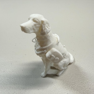 May include: A white figurine of a dog in a seated position, wearing a harness with a rectangular pack on its side. The dog has detailed fur and a collar. The figurine is on a light-colored surface.