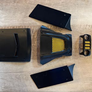 May include: Five black plastic pieces for a car, including a grill with a yellow honeycomb pattern, two side panels, and two smaller panels.