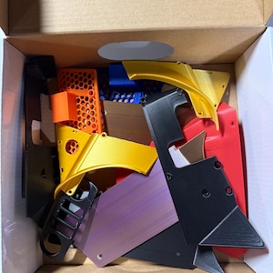 May include: A box filled with various 3D printed parts in different colors, including black, red, orange, gold, purple, and blue. The parts are made of plastic and have a variety of shapes and sizes.