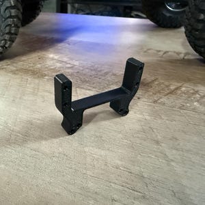 May include: A black plastic 3D printed part with two vertical supports and a horizontal connecting piece. The part is designed to be used as a mounting bracket.