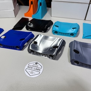 May include: A collection of 3D printed car parts in various colours, including blue, silver, grey, and turquoise. The parts are arranged on a white surface. A sticker with the text "We Print Cool Stuff ROC LABS Est 2023" is visible in the bottom right corner of the image.