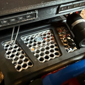 May include: Close-up of a black metal chassis with a honeycomb-shaped metal grill. The chassis has a small, round, gray button with a "G" printed on it. There are also wires and a black electrical component visible.