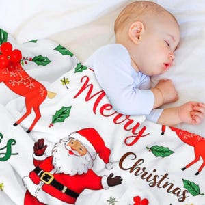 May include: A sleeping baby lies under a white Christmas blanket. The blanket is decorated with a red Santa Claus, reindeer, holly, and the words "Merry Christmas" in red and brown. The baby wears a light blue onesie.