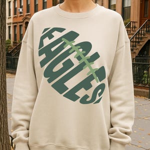 Op de afbeelding: Zandkleurige sweatshirt met een groen voetbalontwerp en het woord "EAGLES" in een gestileerd lettertype. Het ontwerp is gecentreerd op de voorkant. De sweatshirt heeft lange mouwen en een ronde hals.