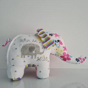 May include: A white plush elephant toy with a floral print on one side and a grey elephant applique with the text "Born in 2020" on the other side. The elephant has a striped ear and a black button eye.
