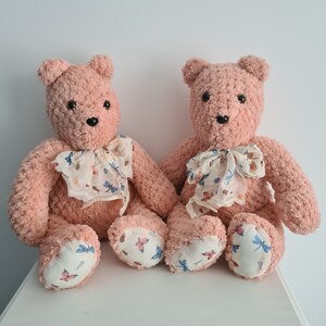 May include: Two plush pink teddy bears with floral patterned scarves. The bears are sitting side by side and are looking at the camera.
