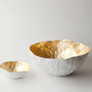 May include: Two decorative bowls. The larger bowl is white with a textured exterior and a gold interior. The smaller bowl has a similar design. Both bowls are on a white background. The bowls are likely for decorative purposes.