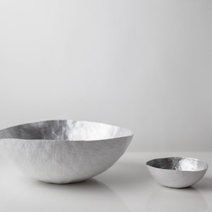 May include: Two silver-colored bowls of different sizes are displayed against a white background. The larger bowl has an irregular shape, while the smaller bowl is round. Both bowls have a textured surface, giving them a unique appearance.
