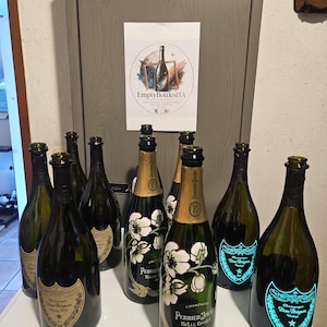 Może przedstawiać: Kolekcja pustych szklanych butelek po szampanie, w tym kilka butelek Dom Pérignon z ciemnozielonego szkła oraz butelki Perrier-Jouët Belle Epoque z białymi motywami kwiatowymi. Widoczny jest znak z napisem EmptyBottlesITA.