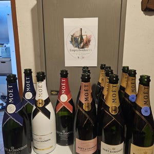 Può includere: Una collezione di bottiglie di champagne Moët & Chandon vuote in vari stili, incluse etichette nere, bianche e rosate. Le bottiglie presentano nastri sul collo e marchi distintivi. Un cartello con il testo EmptyBottlesITA è esposto dietro.