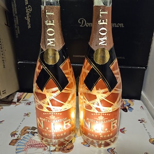 Może przedstawiać: Dwie butelki szampana Moët & Chandon N.I.R Nectar Impérial Rosé Dry z podświetlanym wnętrzem. Butelki mają miedziane, metaliczne wykończenie, czarne etykiety na szyjkach ze złotymi emblematami oraz biały napis N.I.R. na przedniej stronie.