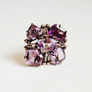 May include: A ring featuring a cluster of five faceted purple amethyst gemstones set in a metallic frame. The stones are arranged in a square-like formation with small metallic spheres interspersed between the individual purple crystals.