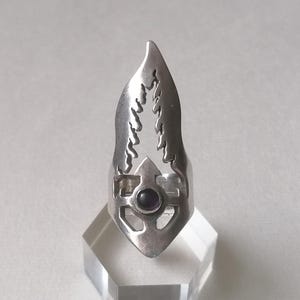 May include: A silver-toned ring with a unique design. The ring features a central purple gemstone and an elongated, stylized shape with cut-out details. The ring is displayed on a clear, geometric stand.