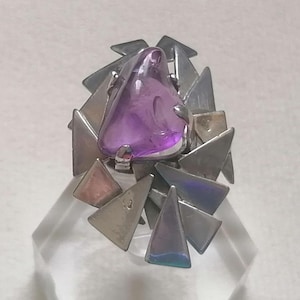 May include: A silver ring with a large, triangular, purple gemstone at its centre. The gemstone is surrounded by numerous silver triangles of varying sizes, creating a unique, geometric design. The ring is set on a clear, angled display.
