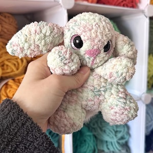 Puede incluir: Un conejito de peluche hecho a mano en colores pastel rosa, verde y blanco. El conejito tiene grandes ojos negros y una nariz rosa. El material suave y texturizado le da un aspecto tierno. El fondo muestra estantes llenos de hilo colorido.