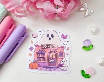May include: A die-cut sticker featuring a pink pumpkin-shaped bookstore with a ghost draped over the roof and another small ghost reading a book in front. The sign reads BOOKSTORE and OPEN. It is surrounded by pink flowers, markers, and small charms.