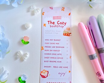 May include: A rectangular paper bookmark titled The Cozy bookshop featuring a list of items including a wool knit blanket, fuzzy socks, and a pumpkin spice latte. The design includes autumn leaves, a barcode, and the text, Redeemable when it smells like cinnamon.