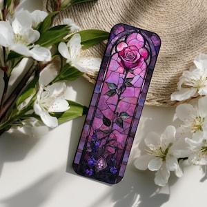 May include: A rectangular bookmark featuring a stained-glass design of a pink rose with green leaves and purple accents. The bookmark is set against a white surface with white flowers and a woven background.