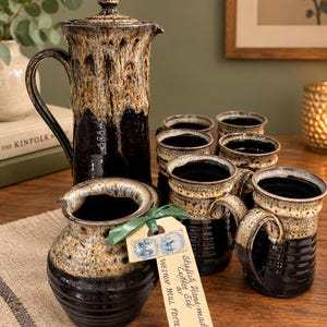 May include: A ceramic coffee set featuring a tall lidded pitcher, a creamer, and four mugs. Each piece has a glossy black base with a textured, dripped cream and brown glaze on the upper portion. A tag reads: Stylish Hand-made Coffee Set by WESTON MILL POTTERY.
